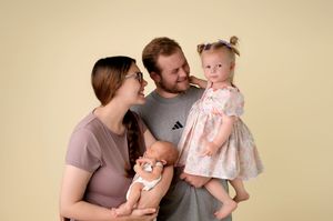 A young family poses for a studio portrait, with mom holding a baby doll and dad lifting their toddler girl in a floral dress.