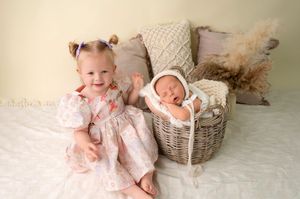 Toddler girl in pink floral dress sits beside a wicker basket holding a sleeping newborn in a white bonnet.
