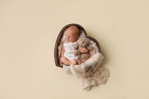 Newborn baby sleeping in a wooden heart bowl, wrapped in cream fabric, holding a stuffed animal toy.