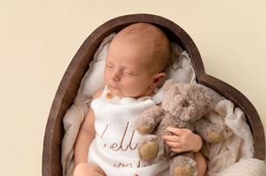 Newborn baby sleeping in heart-shaped wooden bowl wearing white romper, cuddling a soft teddy bear on cream blanket.
