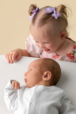 Toddler girl with purple bows smiling at sleeping newborn baby in white crib, sibling love moment.