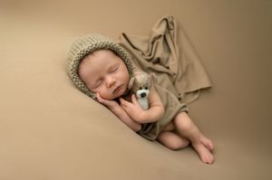 Sleeping newborn baby wearing a knit bonnet, wrapped in a tan blanket, holding a small stuffed animal toy.