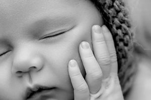 Black and white close-up of a sleeping newborn baby wearing a knit hat with a gentle hand touching its cheek.