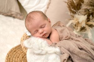 Peaceful newborn baby sleeping in a wicker basket wrapped in a soft beige blanket with white fluffy padding.