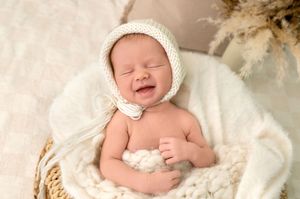 Smiling newborn baby wearing a cream knit bonnet, nestled in a cozy white blanket inside a wicker basket.