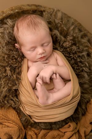 Sleeping newborn baby wrapped in golden swaddle cloth, nestled in a brown fluffy bowl for a professional photo.
