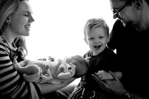 Black and white photo of a joyful family with parents holding a newborn baby while toddler laughs happily.