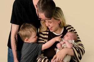 Family portrait with parents and two children, including a newborn baby, smiling together against a beige background.