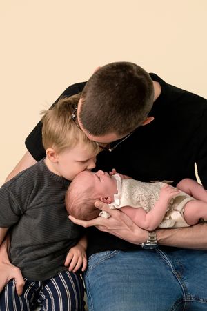 Father holds newborn baby while young sibling leans in for a gentle kiss in a heartwarming family portrait.