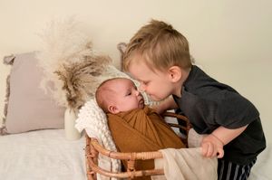 Toddler boy in black shirt kissing newborn baby wrapped in rust swaddle in wicker bassinet.