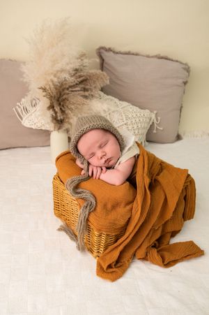 Sleeping newborn baby in knit bonnet nestled in wicker basket with burnt orange blanket and boho decor.