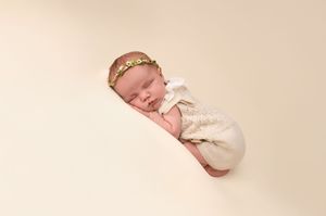 Sleeping newborn baby swaddled in cream wrap with floral headband on soft beige background.