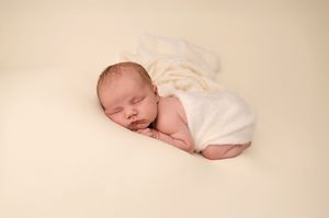 Peaceful newborn baby sleeping wrapped in a soft white blanket on a cream background.