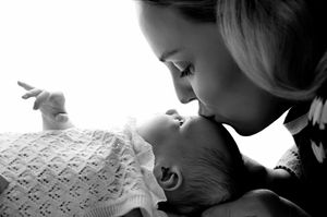 Black and white photo of a mother tenderly kissing her newborn baby lying on a soft blanket.