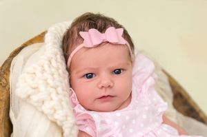 Newborn baby girl in pink polka dot outfit and pink bow headband resting in a basket with white knit blanket.