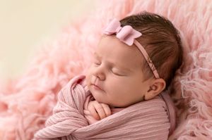 Sleeping newborn baby girl wrapped in pink swaddle with bow headband on fluffy pink blanket.