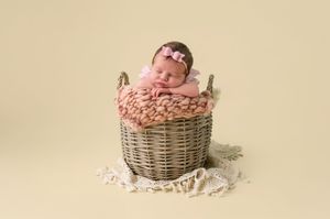 Newborn baby sleeping peacefully in a wicker basket with pink knit blanket and bow headband on cream background.
