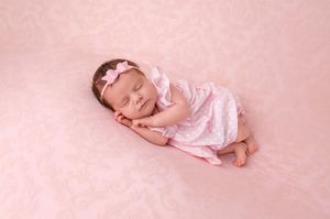 Sleeping newborn baby girl in pink dress and bow headband on pink floral blanket.