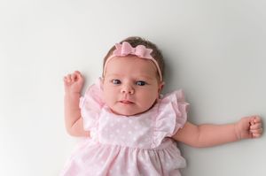 Newborn baby girl in pink polka dot dress and bow headband lying on white background with arms raised.