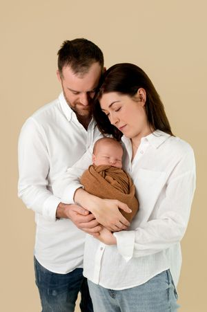 A couple in white shirts tenderly holds their sleeping newborn wrapped in a brown swaddle against a beige background.