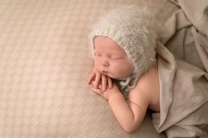 Sleeping newborn baby wearing a soft knit bonnet, posed on a beige textured blanket in a peaceful studio setting.