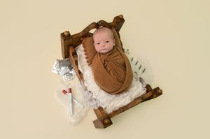 Newborn baby swaddled in brown wrap sleeping in a miniature wooden bed with white fur, on a cream background.