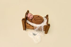 Smiling newborn baby wrapped in brown swaddle sleeping on a miniature wooden bed with white lace blanket.