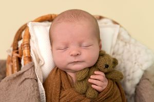 Sleeping newborn baby wrapped in brown knit swaddle holding a small teddy bear in a wicker basket.
