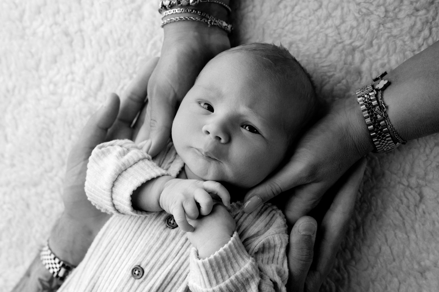 Black and white newborn photo of a baby lying on a soft blanket, cradled gently by two adult hands.