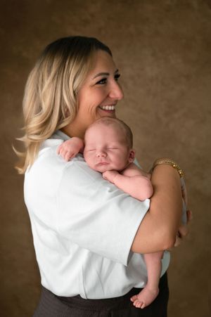 Smiling blonde woman in white blouse holding sleeping newborn baby against her shoulder in studio portrait.