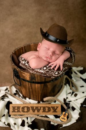 Newborn baby sleeping in wooden bucket wearing tiny cowboy hat with bandana and Howdy sign, western themed photo.