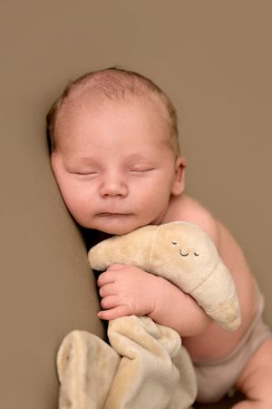 Peaceful newborn baby sleeping while cuddling a soft beige stuffed animal toy against a warm brown background.