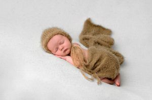 Newborn baby sleeping peacefully wrapped in a soft brown knit blanket and matching bonnet on white backdrop.