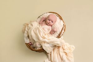 Newborn baby wrapped in cream fabric resting in a wicker basket on a soft beige background.