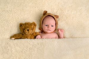Newborn baby wearing a brown bear bonnet lying on a cream blanket next to a matching teddy bear.