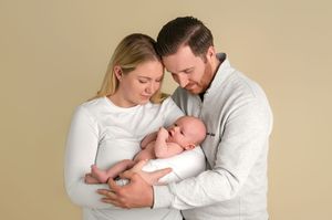 A couple in white tops tenderly gazes at their newborn baby against a neutral beige background.