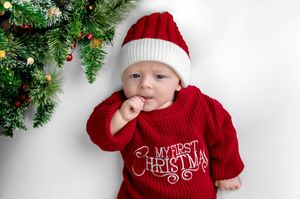 Baby in red My First Christmas sweater and Santa hat lying next to a decorated Christmas tree.