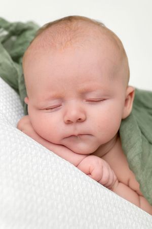Peaceful newborn baby sleeping with chin resting on hand, wrapped in soft green blanket on white background.