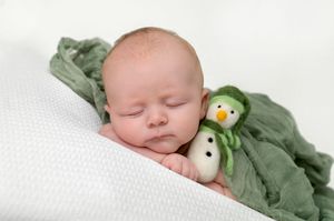 Sleeping newborn baby wrapped in green cloth cuddling a small snowman stuffed toy on white background.