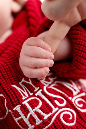 A newborn baby hand gently gripping an adult finger on a red Merry Christmas knit blanket.