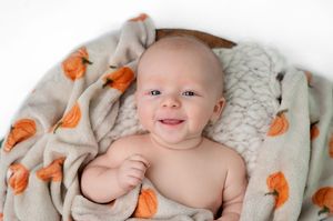 Smiling baby wrapped in a cozy beige blanket with orange pumpkin print, lying in a round wooden bowl.
