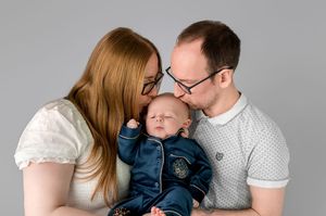 Parents kissing their newborn baby on the cheeks in a professional studio portrait on a gray background.