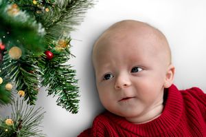 Baby in red sweater lying next to decorated Christmas tree branches with bokeh lights.
