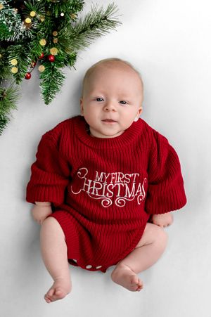 Baby in red My First Christmas sweater romper lying next to decorated Christmas tree branches.