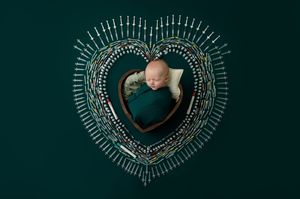 Newborn baby sleeping in teal wrap inside decorative heart-shaped frame on green background.
