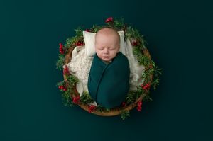Newborn baby wrapped in green swaddle sleeping in a wooden bowl with a festive Christmas wreath for holiday photos.
