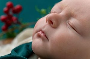 Sleeping newborn baby with delicate features, dressed in green, with festive holly berries in the background.