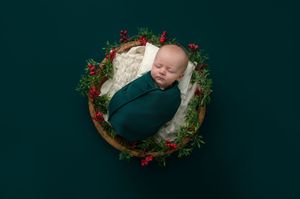 Newborn baby swaddled in green wrap, sleeping in a wooden bowl decorated with holly berries and greenery.