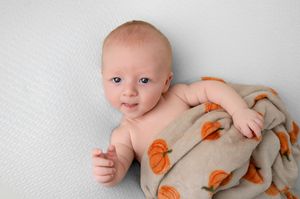 Cute baby lying on white surface wrapped in a tan blanket with orange pumpkin designs, smiling softly.
