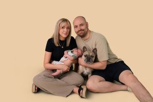 A couple sits on a beige backdrop holding a newborn baby and a French Bulldog for a family portrait photo.